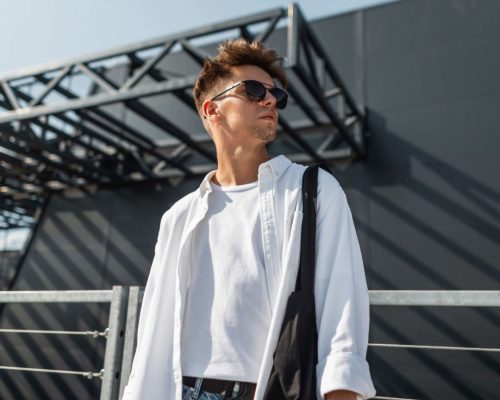 attractive-young-man-stylish-white-shirt-fashionable-sunglasses-trendy-jeans-with-cloth-black-bag-posing-sunny-day-near-gray-building-outdoors-american-guy-model-youth-summer-style