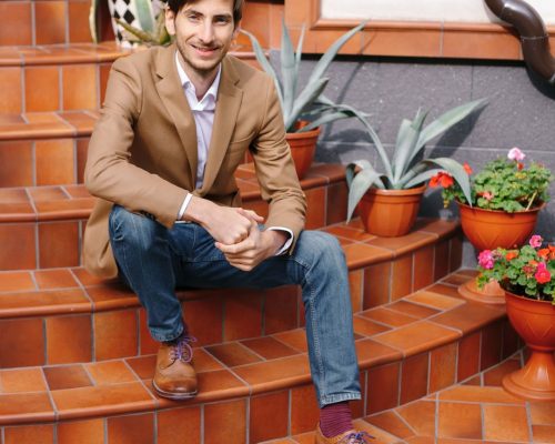smiling-young-stylish-man-sitting-outdoors-vintage-circular-stairs